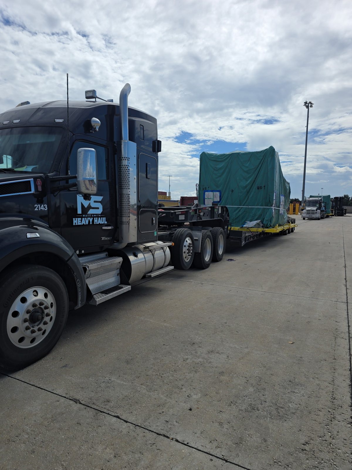MS Heavy Haul Kenworth T880, truck 2143, with a green-tarped oversize load on a multi-axle lowboy, staged on a terminal lot in Mobile, Alabama