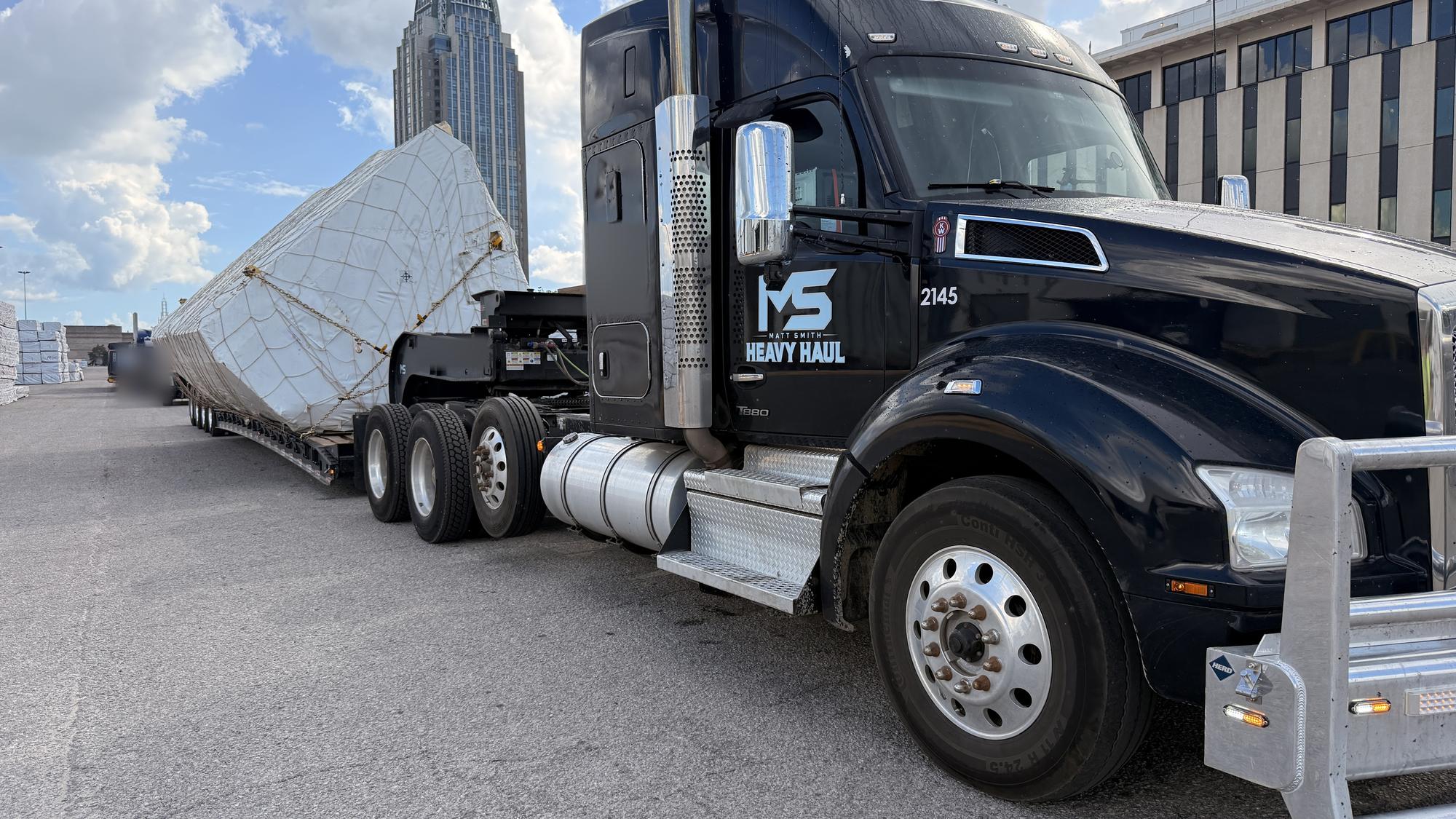 MS Heavy Haul Kenworth with a shrink-wrapped oversize load on a multi-axle lowboy, downtown Mobile, Alabama skyline behind