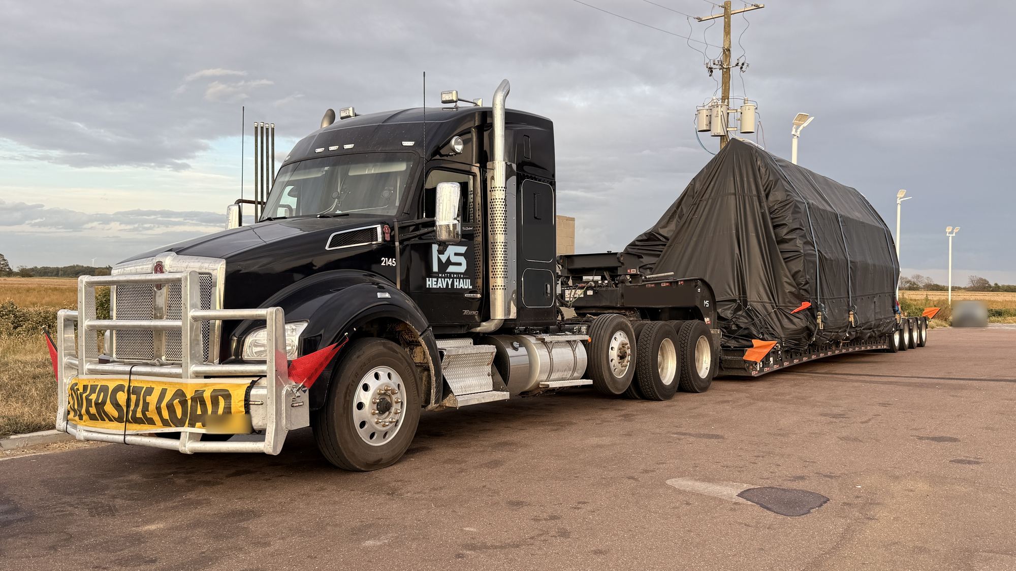 MS Heavy Haul Kenworth 2145 with an oversize load banner and a black-tarped load on a multi-axle lowboy, parked beside a field