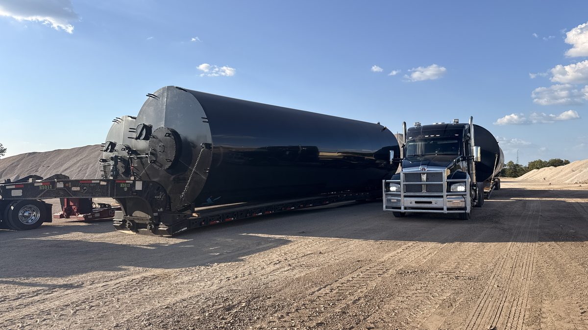MS Heavy Haul black Kenworth with a long black tank loaded on an extended stretch RGN in a sand yard, with a second tank load on another truck behind it