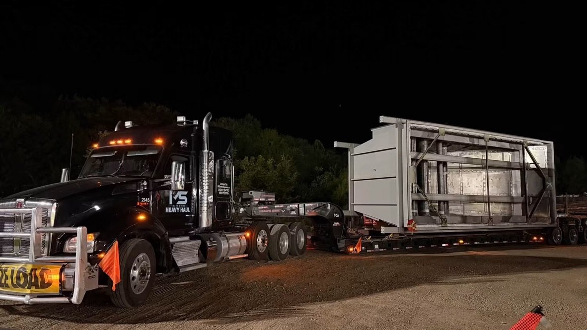 Black Kenworth with oversize load banner hauling a steel structure on a multi-axle lowboy at night